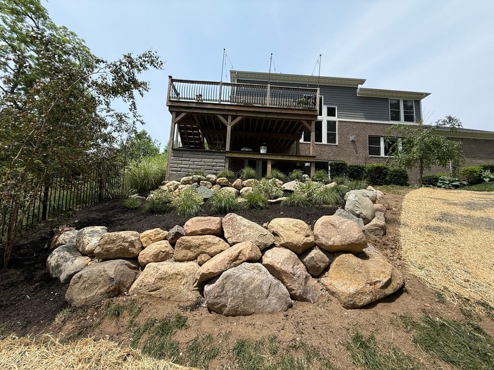 Backyard landscape featuring a stone retaining wall and elevated wooden deck with greenery.