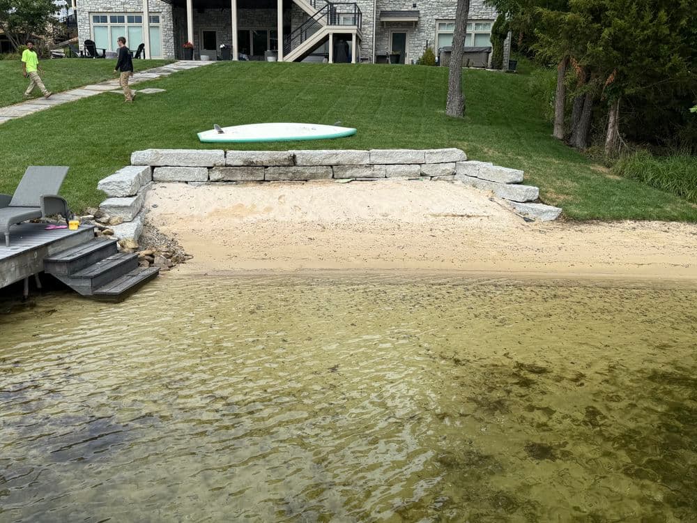 View of a sandy beach area with stone wall, clear water, and paddleboard at a lakeside property.