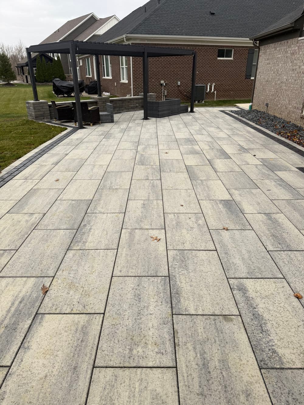 Modern patio with gray pavers, surrounded by landscaped lawn and a pergola in the background.